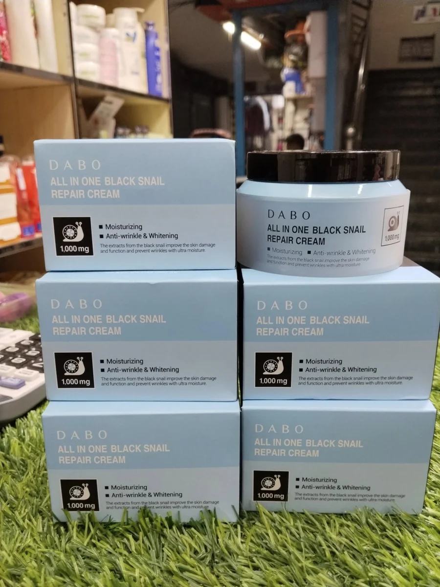 Multiple boxes and a jar of DABO All In One Black Snail Repair Cream arranged on green artificial grass in a shop setting.