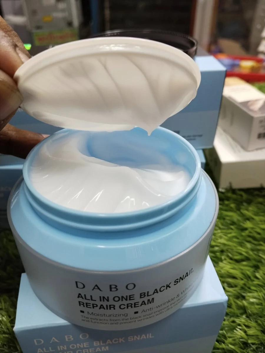 Multiple boxes and a jar of DABO All In One Black Snail Repair Cream arranged on green artificial grass in a shop setting.