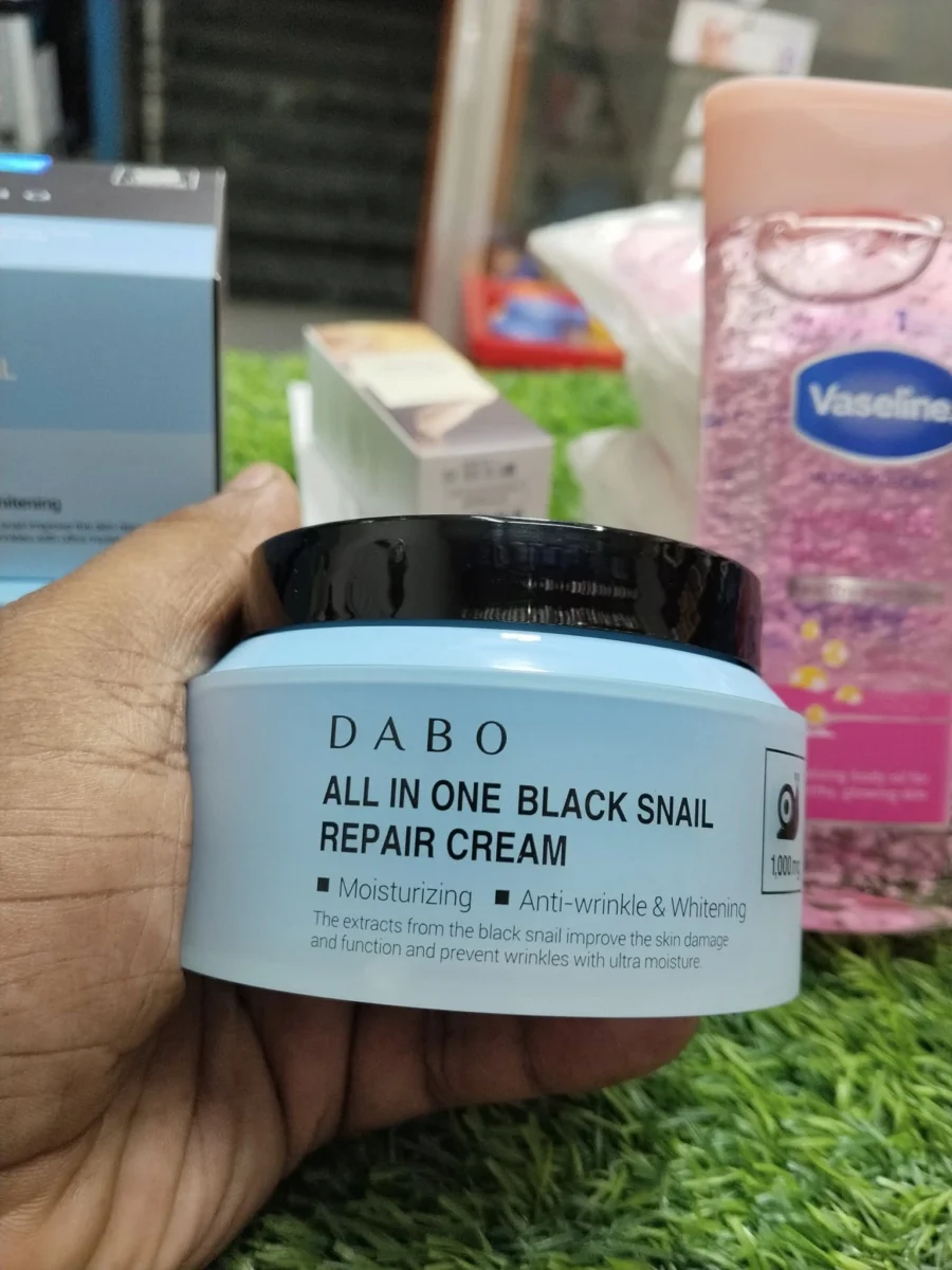 Multiple boxes and a jar of DABO All In One Black Snail Repair Cream arranged on green artificial grass in a shop setting.